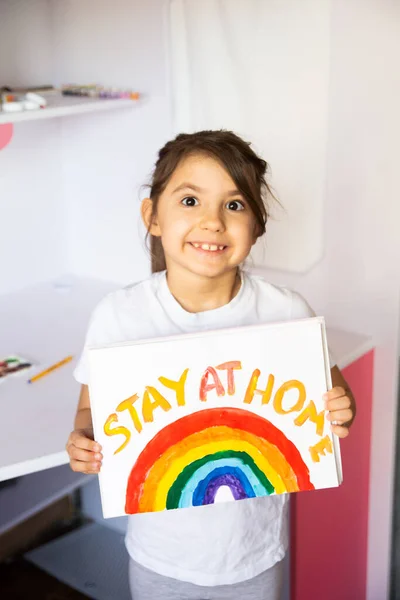 Stay at home due coronavirus pandemic concept. Little girl paints a rainbow on stay at home poster. Chase the rainbow flashmob. Positive activities during quarantine and staying at home. COVID - 2019.