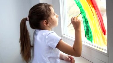 Evde Covid-19 karantinası sırasında küçük bir kız pencereye gökkuşağı çiziyor. Gökkuşağı flaş çetesini kovala. Umarım her şey yolunda gider.