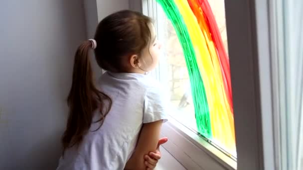 Een klein meisje kijkt uit het raam met een geschilderde regenboog tijdens coronavirus quarantaine thuis. Achtervolg de regenboog flitsmeute en blijf thuis concept. — Stockvideo