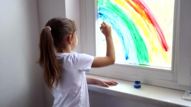 Küçük beyaz kız evdeki koronavirüs karantinası sırasında pencereye gökkuşağı çiziyor. Gökkuşağı flaş kalabalığını kovala