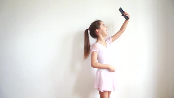 Piccola ragazza caucasica carina che guarda nel telefono e ride, prende selfie e posa per le foto, chatta con gli amici online. Comunicazione online e concetto di blogging . — Video Stock