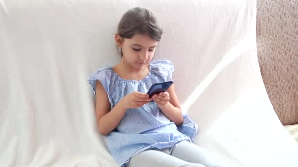 Kíváncsi aranyos lány óvodás gyermek segítségével egy digitális technológia eszköz néz telefon. Játszik a telefonban, egyedül ül a kanapén. A karantén ideje alatt otthon lakó gyermekek COVID-19 — Stock videók
