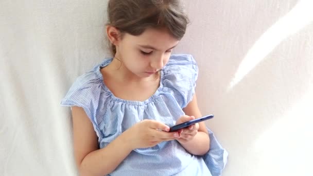 Fermer à l'aide d'un smartphone. Joue dans le téléphone assis sur le canapé seul. Confort et un moment agréable à la maison. Enfants restant à la maison et étudiant à distance pendant la quarantaine COVID-19 — Video