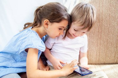 Erkek ve kız kardeşler tabletle oynuyorlar. Çocuklar evde iyi vakit geçirsin. Çocuklar kanepede oturup tabletle oynuyorlar. Çocuklar cep telefonuyla oyun oynuyorlar. Ev konseptinde kal