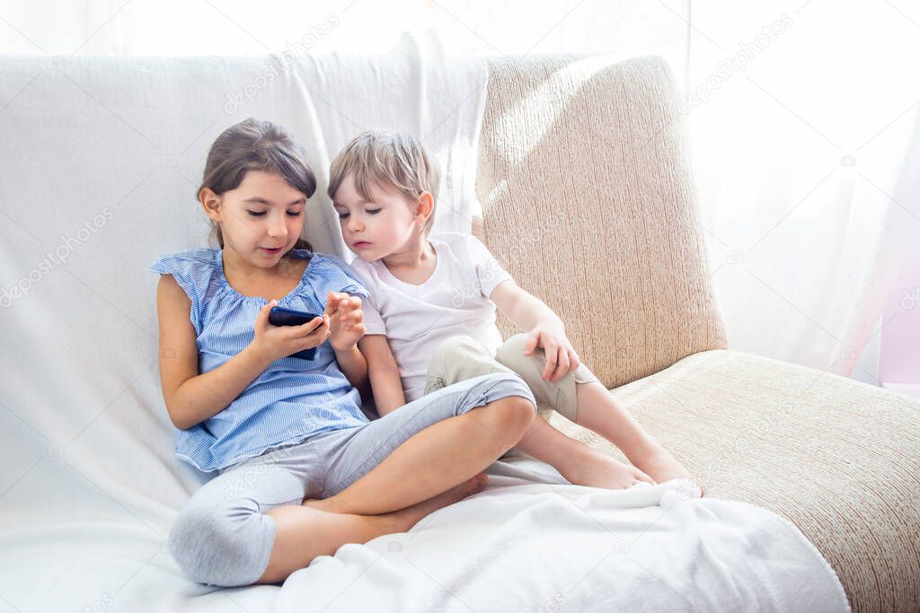 Children at home sitting on sofa, playing with tablet. Cute siblings brother and sister have a good time at home. Kids play a game on a cell phone. Stay at home during quarantine COVID-19 concept