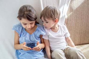 Çocuklar ev kavramında iyi vakit geçiriyor. Yakın küçük kardeş tabletle oynuyor. Çocuklar kanepede oturup tabletle oynuyorlar. Çocuklar cep telefonuyla oyun oynuyorlar.