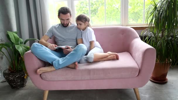 Concepto del día de los padres. Papá le está leyendo un libro a una linda niña. Papá y su hija se sonríen el uno al otro y disfrutan quedarse en casa durante el mal tiempo . — Vídeos de Stock