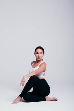 Kız yoga için poz veriyor, siyah taytlarla ısınıyor ve beyaz arka planda beyaz bir tişört giyiyor.