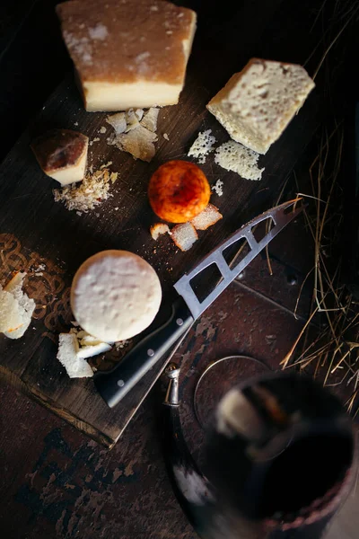 Diferentes quesos se cortan en una tabla de madera junto a un cuchillo para cortar queso en una caja vieja con heno y una botella — Foto de Stock