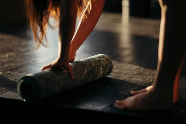 Una chica enrolla una alfombra con las manos y se para en los dedos del pie en el suelo para hacer yoga en un gimnasio o en casa en un clima soleado — Foto de Stock