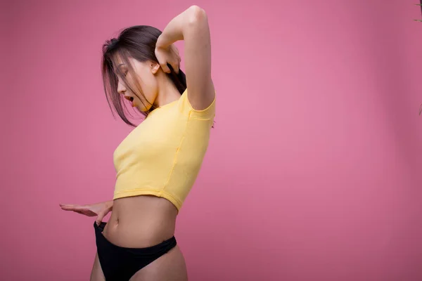 Beautiful Asian woman posing and showing how thin she pulls black panties in f yellow t-shirt on a pink background — 스톡 사진