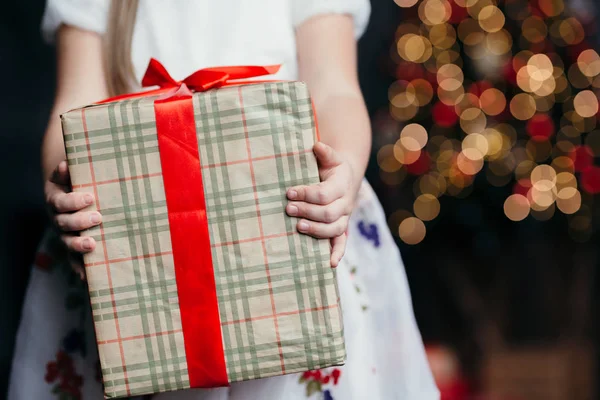 Niña con un vestido blanco sostiene un regalo de Navidad de papel artesanal con una cinta roja sobre un fondo del árbol de Navidad con guirnaldas hermosas luces —  Fotos de Stock