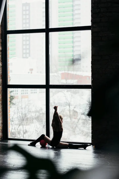 Die Turnerin sitzt auf einem Bindfaden und benutzt den Ball. Silhouette eines Mädchens auf hellem Fensterhintergrund. — Stockfoto