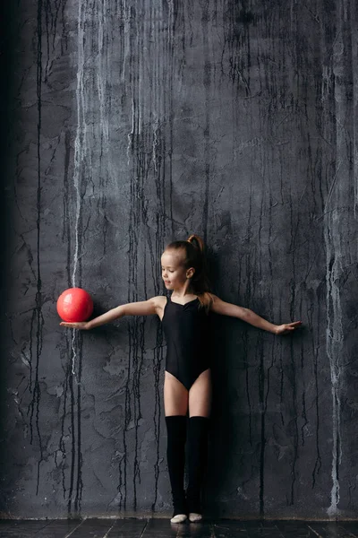 Flicka gymnast poserar med en röd boll. Gymnast klädd i sportkläder med golf. På bakgrunden är en svart strukturerad vägg. — Stockfoto