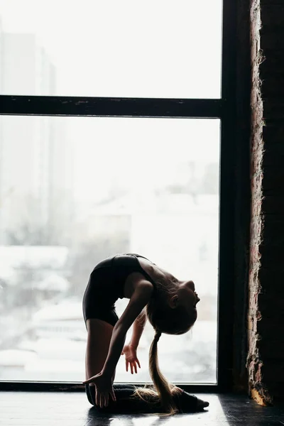 De gymnaste knielt bij het raam. Silhouet van een meisje op een heldere achtergrond. — Stockfoto