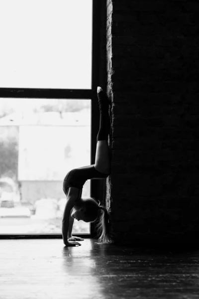 Die Turnerin liegt sich in den Armen. Silhouette eines Mädchens auf einem hellen Fensterhintergrund. Schwarz-Weiß-Foto. — Stockfoto