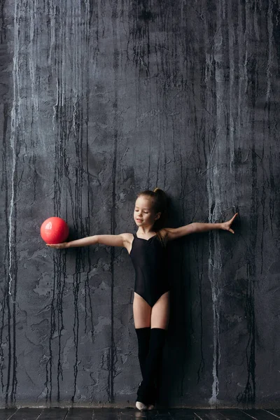Ginnasta ragazza in posa con una palla rossa. Ginnasta vestita in abbigliamento sportivo con golf. Sullo sfondo è un muro nero strutturato . — Foto Stock