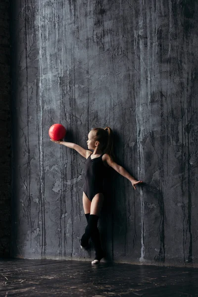 Gymnaste fille posant avec une boule rouge. Gymnaste vêtu de vêtements de sport avec golf. Sur le fond est un mur texturé noir . — Photo