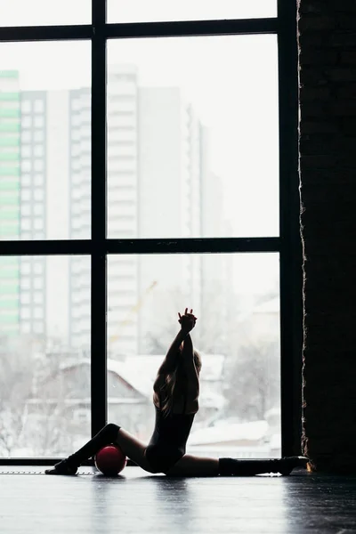 Die Turnerin sitzt auf einem Bindfaden und benutzt den Ball. Silhouette eines Mädchens auf hellem Fensterhintergrund. — Stockfoto