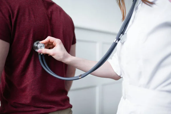 Close-up um médico em um casaco branco examina um paciente do sexo masculino com um fonendoscópio para diagnosticar coronavírus — Fotografia de Stock