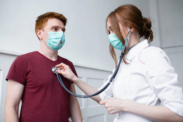 O paciente ruivo experimenta ataques de tosse e dor de cabeça e é examinado por uma linda médica em uma máscara. — Fotografia de Stock