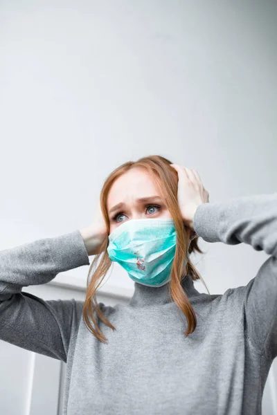 Menina de cabelos vermelhos em uma máscara azul com manchas de sangue olha pensativo enquanto espera por uma consulta de médicos — Fotografia de Stock