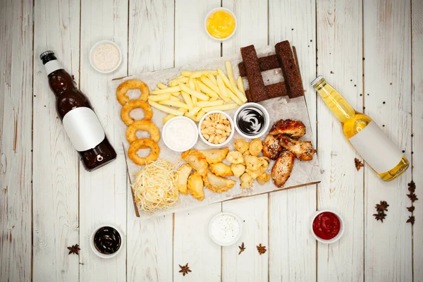 Ahşap bir masanın üzerinde abur cubur, sos ve bira şişeleri var. Beyaz tabakta patates, kızarmış ekmek, tavuk ve soslar var..