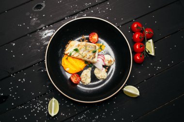Siyah tabakta taze sebzelerle servis edilen ızgara balık. Siyah ahşap bir masanın üzerinde kiraz domatesleri ve limon dilimleri uzanıyor..