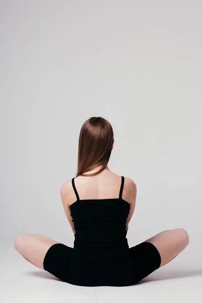 Молода дівчина в чорному спортивному одязі сидить з нею спиною до камери на білому тлі і медитує — стокове фото
