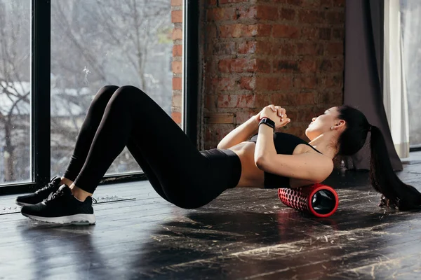 Una chica en ropa deportiva hace ejercicios para fortalecer sus músculos de la espalda usando un cilindro deportivo. Entrenamiento matutino en el gimnasio antes del trabajo —  Fotos de Stock