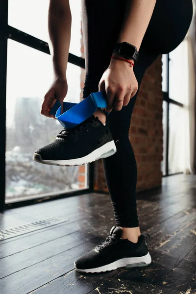 Yakın plan. Kız bacak kasları üzerinde egzersiz yapmak için mavi lastik bant çekiyor. Siyah elbiseler ve spor ayakkabılar.. — Stok fotoğraf