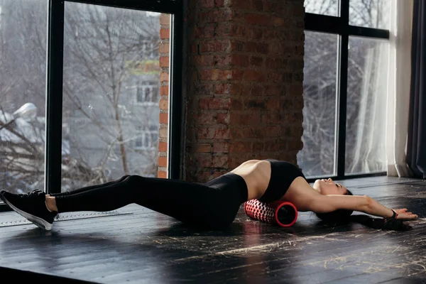 Mujer joven en el gimnasio en el entrenamiento. El atleta hace ejercicios con un cilindro deportivo tirado en el suelo. Entrenamiento para fortalecer los músculos de la espalda y los abdominales — Foto de Stock