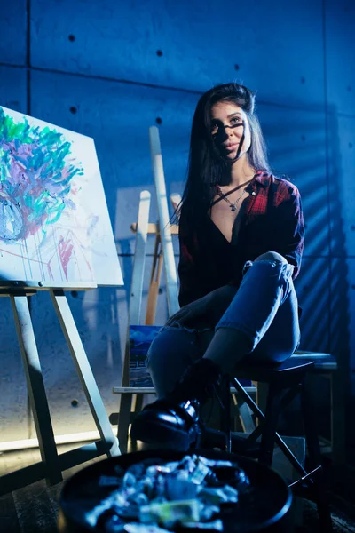 Meisje kunstenaar zittend op een stoel in een donker atelier in de buurt van geschilderd abstract acryl schilderij. — Stockfoto