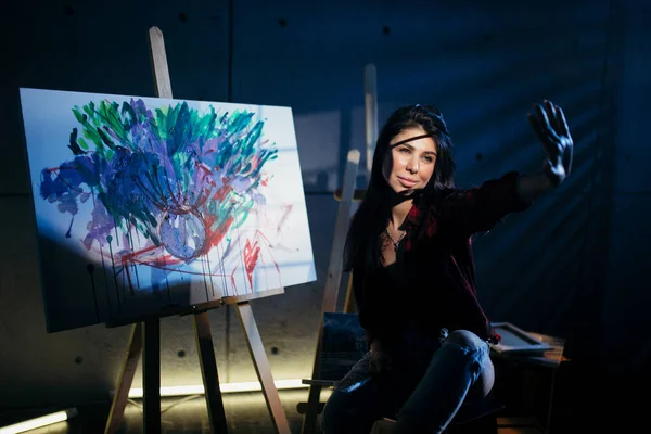 Beautiful brunette girl artist sitting in a dark Studio near the easel with painted watercolor abstract painting. — Stock Photo, Image