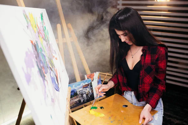 Brunett artist blandar färg på en träpalett nära staffliet och målad med akrylfärg målning. — Stockfoto