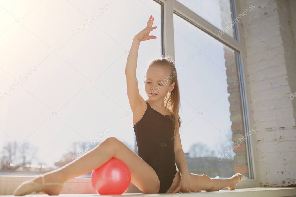 Talented gymnast did the splits and beautifully pulled his hand up. The girl with a red ball on the background of the window.