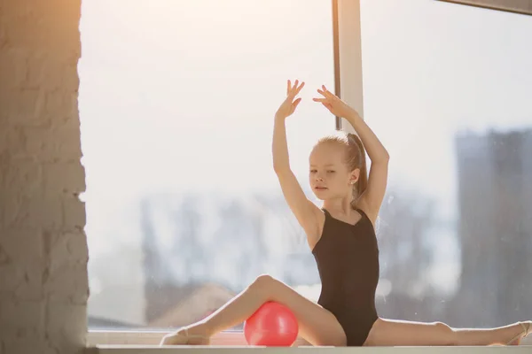 Gymnaste talentueux a fait les fentes et magnifiquement tiré sa main vers le haut. La fille avec une boule rouge sur le fond de la fenêtre . — Photo