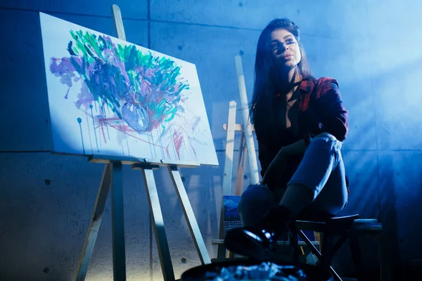 Beautiful brunette girl artist sitting in a dark Studio near the easel with painted watercolor abstract painting. — Stock Photo, Image