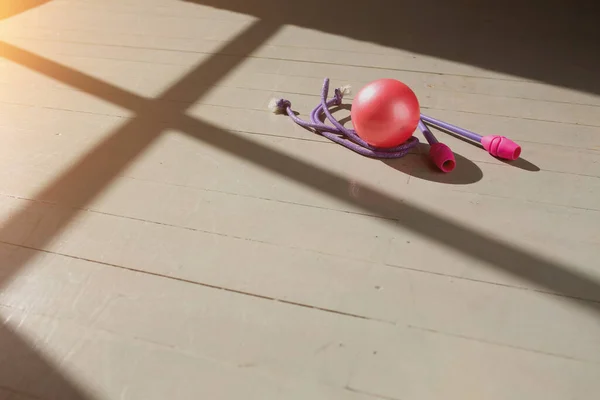 Pink gymnastic wands, pink ball and jump rope lying on the wooden floor in the rays of the rising sun. Bright light and shadow.