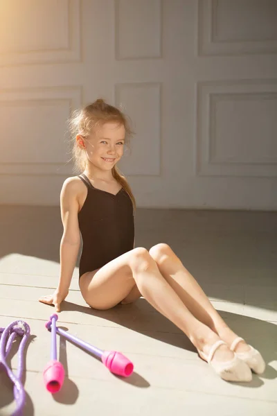 Little girl in a black bathing suit sits beautifully against the window and near are gym ball, jump rope, and pink rods. — Stock Photo, Image