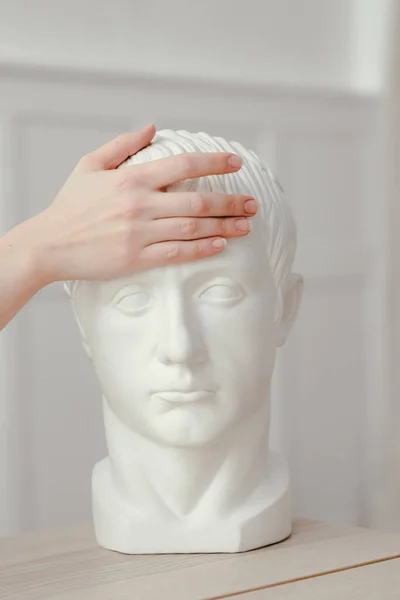 La mano femenina sostiene una cabeza de yeso esculpida detrás de la frente. Medición de temperatura a través del tacto . —  Fotos de Stock