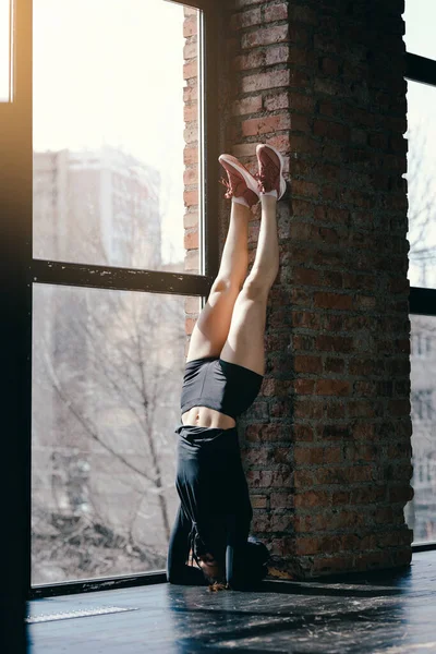 Ο προπονητής Πιλάτες κάνει κατακόρυφο κοντά σε τοίχο από τούβλα. Αθλητικό κορίτσι με μαύρα ρούχα σήκωσε τα πόδια. — Φωτογραφία Αρχείου