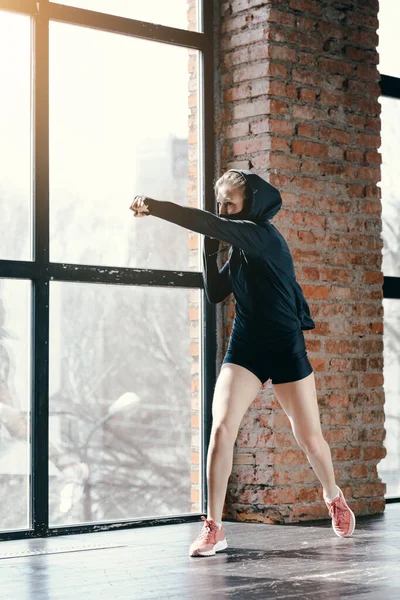 Blondă sport într-o jachetă întunecată îndeplinește lovituri în sala de sport lângă fereastra panoramică în fața competiției . — Fotografie, imagine de stoc