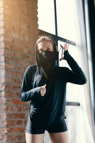 Ένα νεαρό κορίτσι με μαύρη κουκούλα διορθώνει τη μαύρη μάσκα στο πρόσωπο. Πυγμαχία προπονητής πήρε ένα διάλειμμα κατά τη διάρκεια μιας κατ 'οίκον προπόνηση. — Φωτογραφία Αρχείου