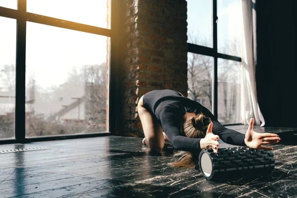 Gimnasta flexible está sentado en el suelo y haciendo curvas hacia adelante en el rodillo de masaje. El yogui dobla el torso a las piernas . — Foto de Stock