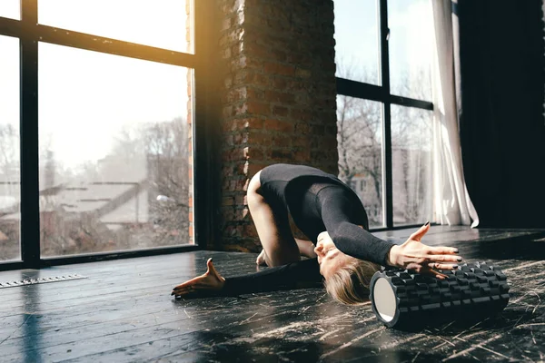 Gimnasta flexibilă este așezată pe podea și face curbe înainte pe role de masaj. Yoghinul îndoaie trunchiul la picioare . — Fotografie, imagine de stoc