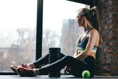 Spor kıyafetli bir kız gergin bir egzersizden sonra stüdyoda yerde oturuyor, yanında bir masaj silindiri ve miyofasyal top var..