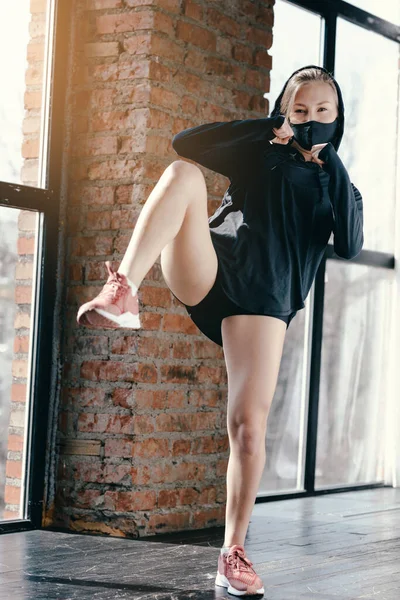 Chica atlética joven en chaqueta negra y máscara cumple patadas durante un estudio de artes marciales con ventanas panorámicas y pared de piedra . —  Fotos de Stock