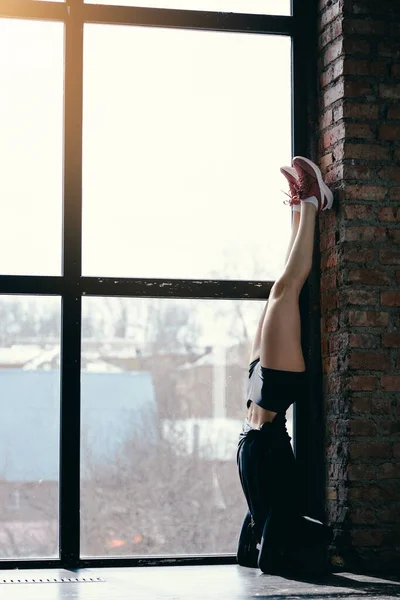 Una esbelta muchacha en pantalones cortos está de pie en sus brazos verticalmente contra la pared. Soporte de mano vertical. Ventana panorámica . — Foto de Stock