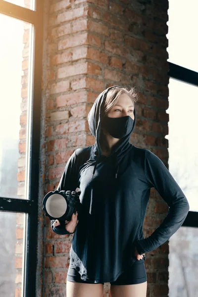 Mladá sportovní dívka v černé masce drží v rukou masážní válec a připravuje se na domácí trénink v podmínkách sebeizolace. — Stock fotografie